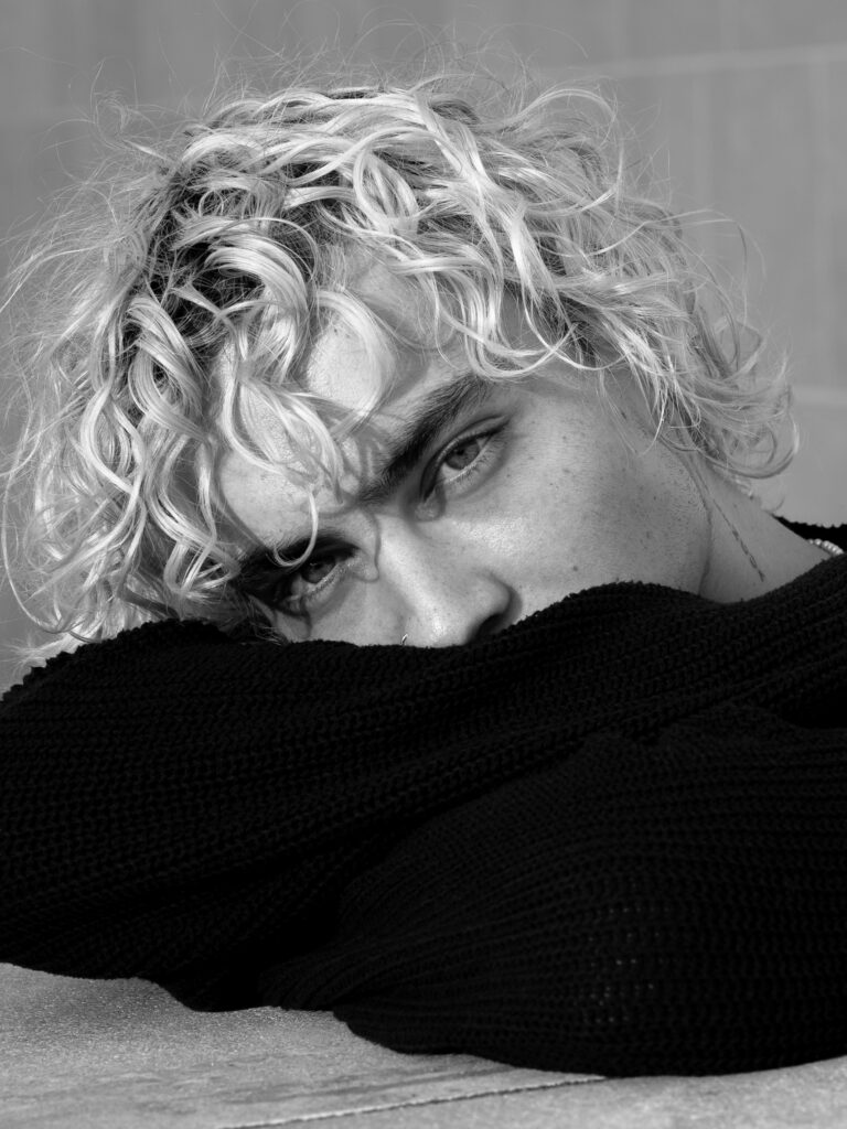 Portrait noir et blanc d’un homme aux cheveux bouclés photographié en lumière naturelle, regard intense vers l’objectif.